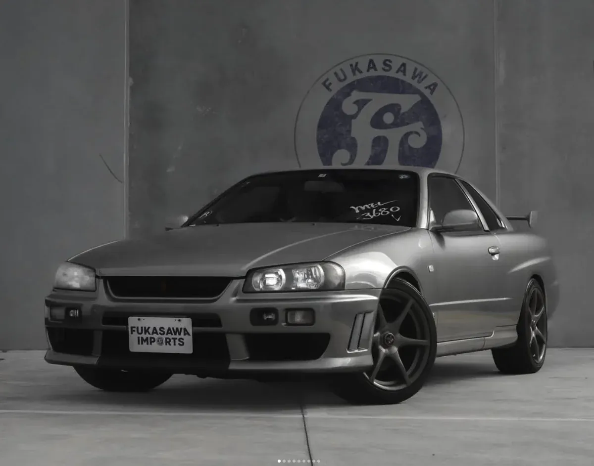 Silver Nissan Skyline R34 coupe front three-quarter view in front of the Fukasawa Car Imports logo wall